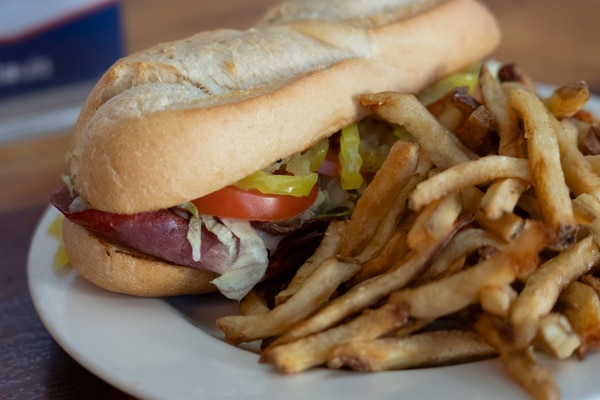 Metal takeout bowl with rice topped by a sunny-side-up egg, sliced sausage and a tomato-onion salad with a small cup of chopped onions and scallions, set on a wooden table beside a woven bamboo tray holding a small potted succulent.