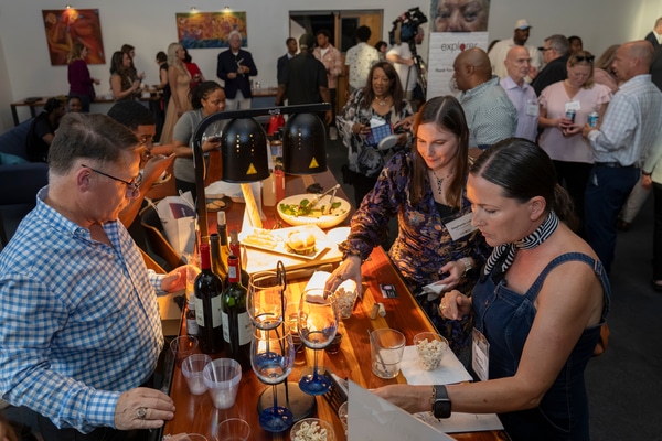 Red brick Victorian-style house with a rounded turret and arched porch behind a low stone wall, an American flag and flowering shrubs at the entrance under an overcast sky.