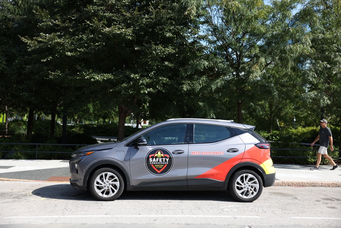 Long sunlit café corridor with floor-to-ceiling windows on the right casting rectangular light patches and a cursive shadow on the floor, a large carved circular wall medallion on the left, and tables with a city street visible beyond.