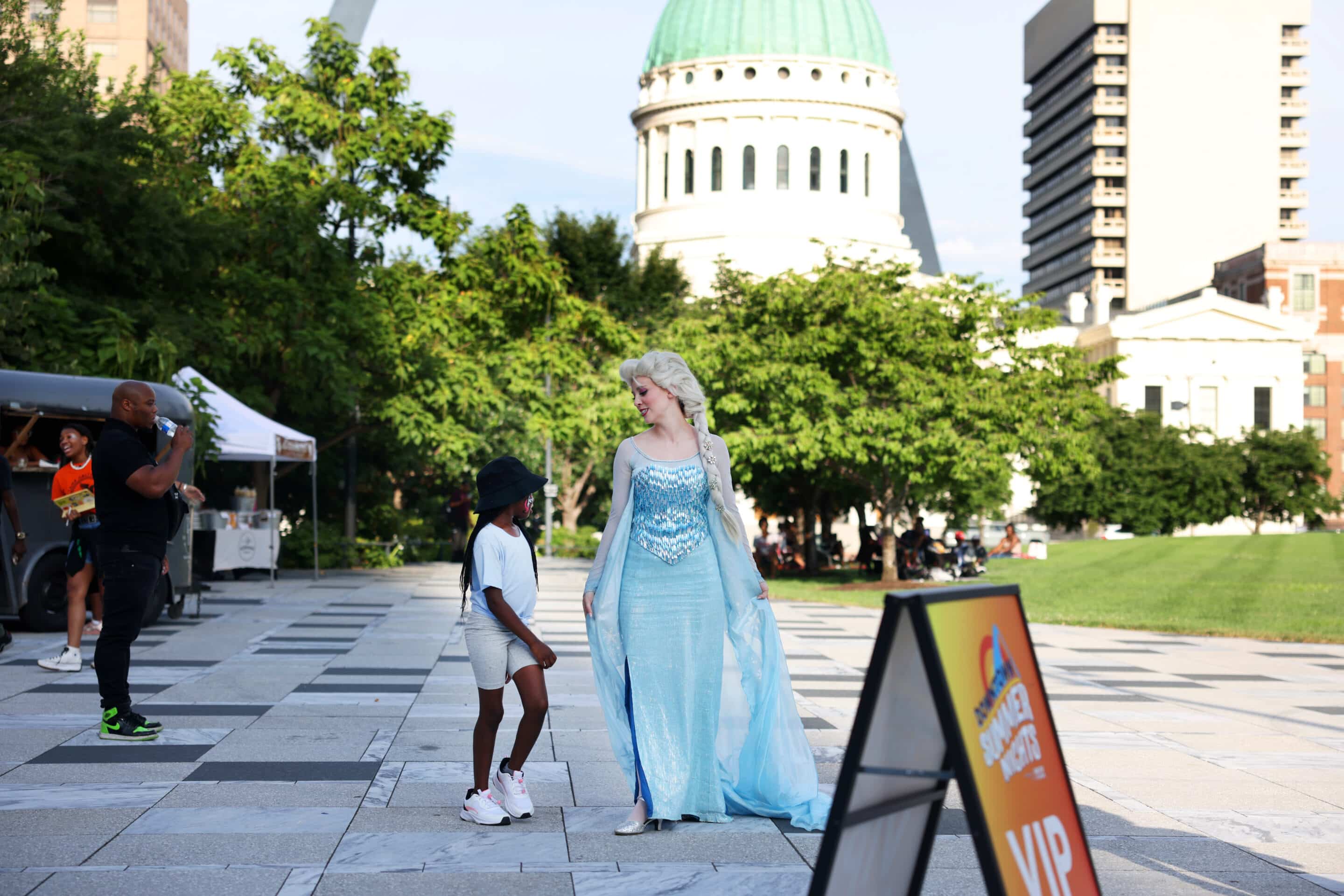 Rooftop swimming pool and sun deck with lounge chairs and a woven egg chair in the foreground, blue water bordered by a Greek-key tile band, and a city skyline under a bright, partly cloudy sky.