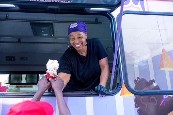Metal takeout bowl with rice topped by a sunny-side-up egg, sliced sausage and a tomato-onion salad with a small cup of chopped onions and scallions, set on a wooden table beside a woven bamboo tray holding a small potted succulent.