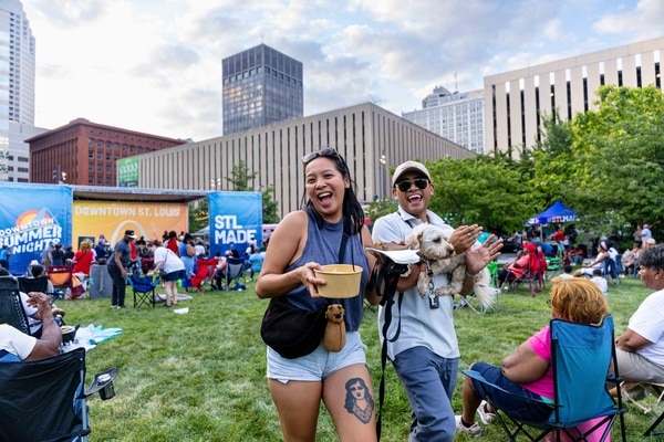 Red brick Victorian-style house with a rounded turret and arched porch behind a low stone wall, an American flag and flowering shrubs at the entrance under an overcast sky.