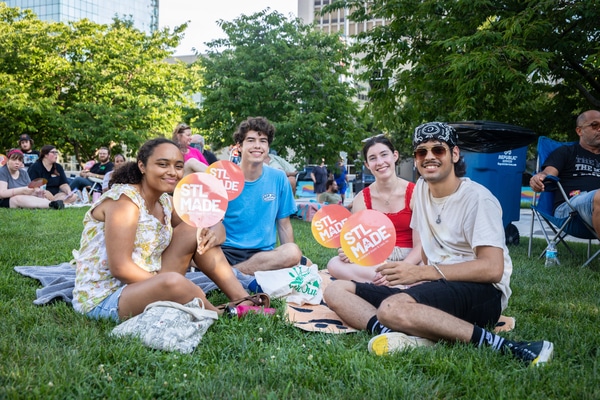Rooftop bar at dusk with hanging string lights, people dining and lounging on curved sofas around a fire pit, a bartender at the bar, and a large arch and bridge on the city skyline.