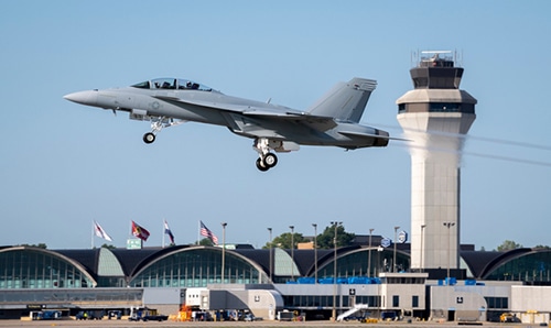 Gray military fighter jet with landing gear down flying over an airport terminal with a control tower in the background.