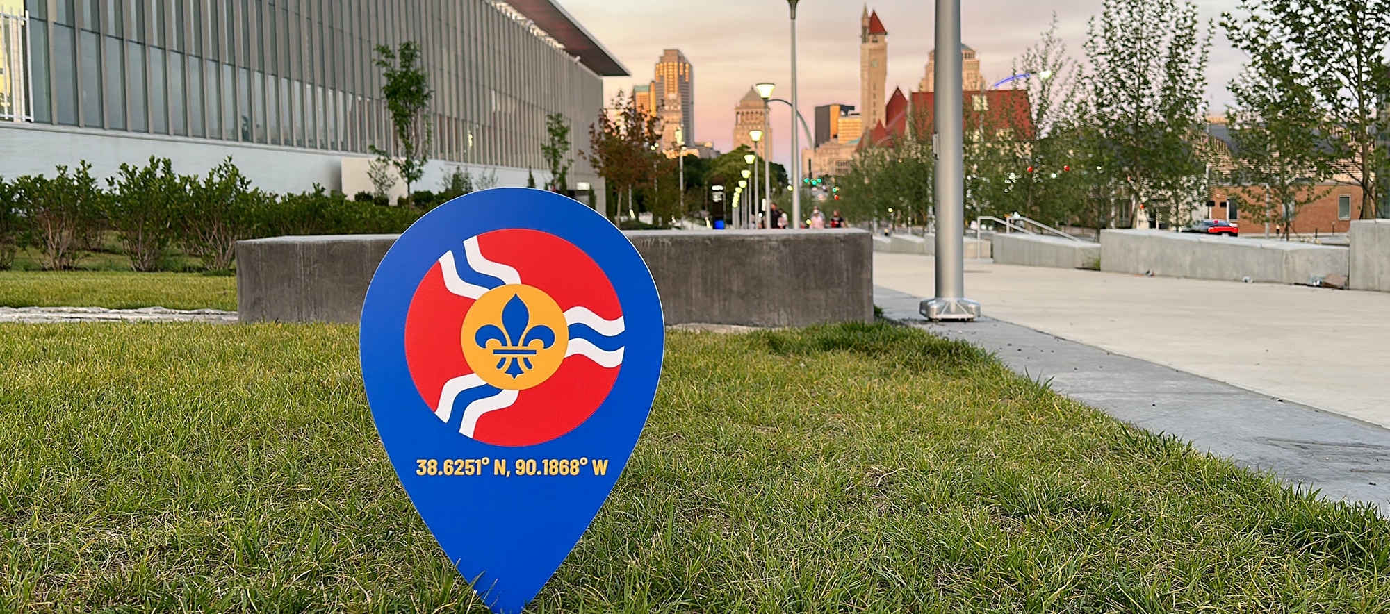 Blue map-pin sign with fleur-de-lis and coordinates 38.6251°N, 90.1868°W in grass by an urban walkway.