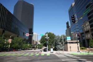 Downtown city intersection on Market Street with bright green-striped crosswalks, glass-clad office towers lining the street, traffic lights, street signs and trees under a clear blue sky.