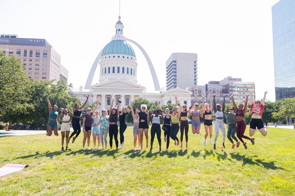 Rooftop swimming pool and sun deck with lounge chairs and a woven egg chair in the foreground, blue water bordered by a Greek-key tile band, and a city skyline under a bright, partly cloudy sky.