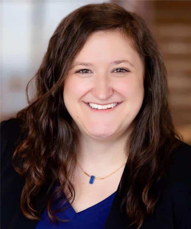 Smiling woman with long wavy brown hair wearing a blue top, black blazer and a blue pendant necklace, blurred background.