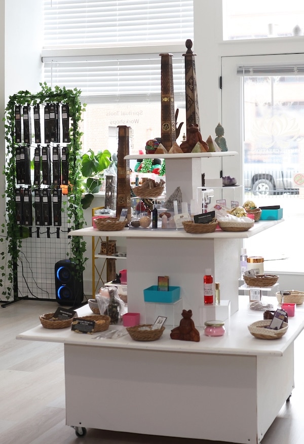 White three-tier display stand in a bright shop holding baskets, small gifts and wooden totems, with a plant and rack nearby.