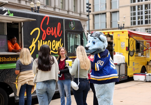 Stainless-steel Gateway Arch towering over a broad green park with a circular fountain and walkways, framing the downtown St. Louis skyline under a bright blue sky.