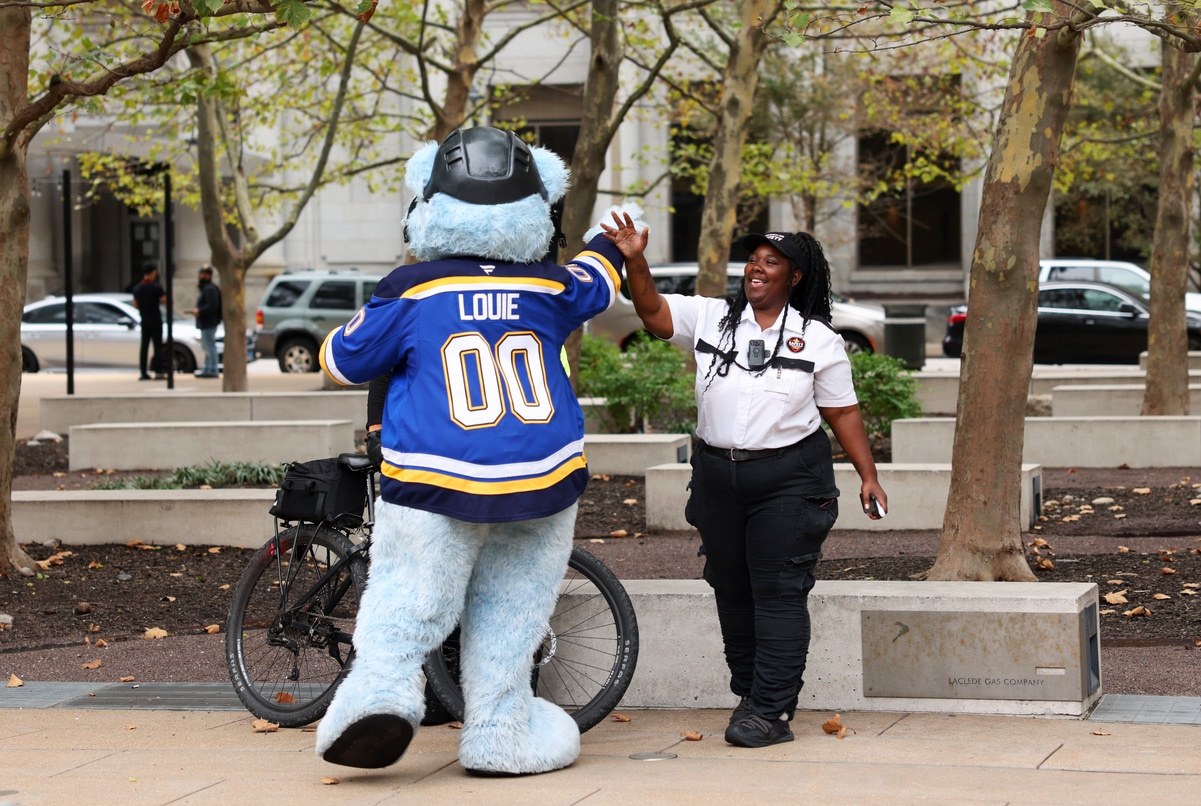 Blue furry hockey mascot Louie wearing a blue No. 00 jersey and helmet high-fives a smiling female security officer in a white shirt and black pants in a tree-lined urban plaza with a bicycle and parked cars behind them.
