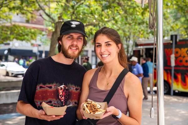 Metal takeout bowl with rice topped by a sunny-side-up egg, sliced sausage and a tomato-onion salad with a small cup of chopped onions and scallions, set on a wooden table beside a woven bamboo tray holding a small potted succulent.