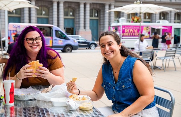 Rooftop bar at dusk with hanging string lights, people dining and lounging on curved sofas around a fire pit, a bartender at the bar, and a large arch and bridge on the city skyline.