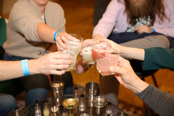 Metal takeout bowl with rice topped by a sunny-side-up egg, sliced sausage and a tomato-onion salad with a small cup of chopped onions and scallions, set on a wooden table beside a woven bamboo tray holding a small potted succulent.