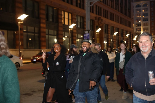 Red brick Victorian-style house with a rounded turret and arched porch behind a low stone wall, an American flag and flowering shrubs at the entrance under an overcast sky.
