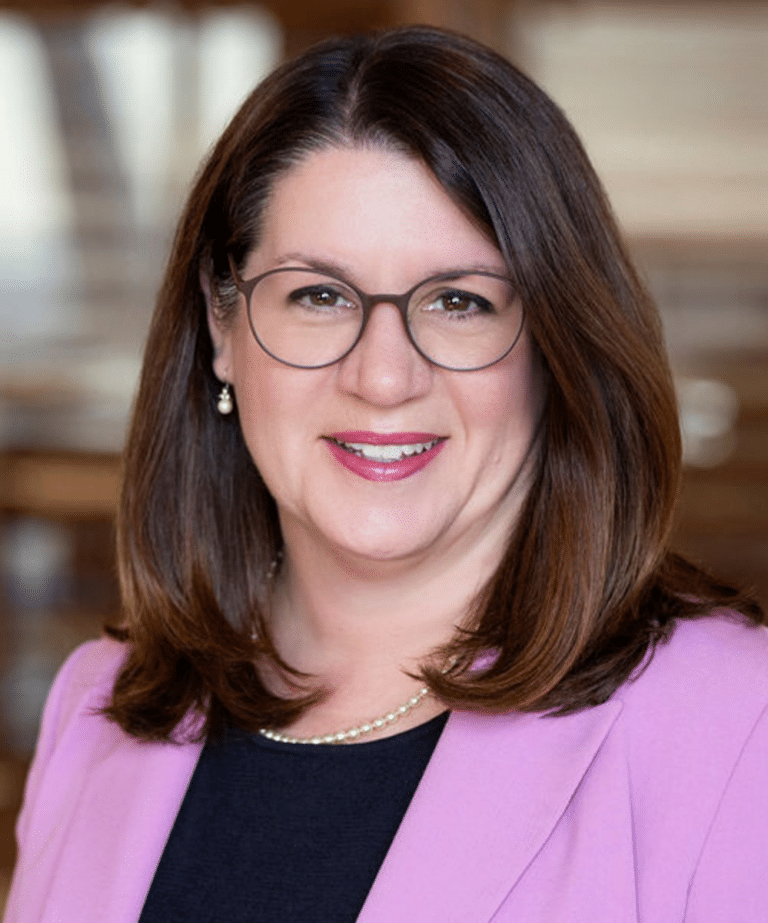 Smiling woman with shoulder-length brown hair, round glasses, pearl necklace and pink blazer against blurred background.