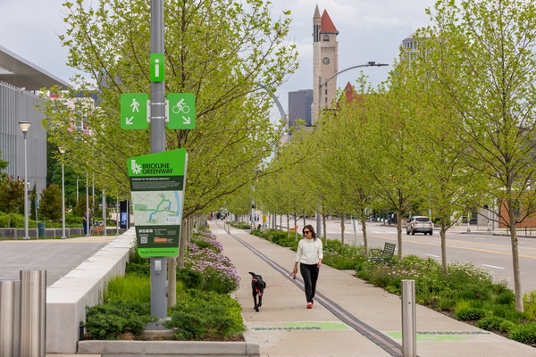 Woman in a white sweater walking a black dog on a leash along a tree-lined urban greenway path with a green 'Brickline Greenway' sign on a pole and a clock tower visible among downtown buildings in the background