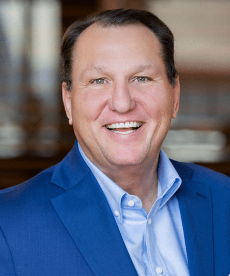 Smiling middle-aged man in a blue suit and light-blue shirt, close-up headshot with a blurred warm-toned background.