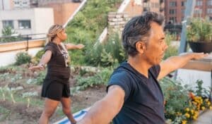 Two people standing on a rooftop garden with arms outstretched practicing yoga, city buildings in the background.