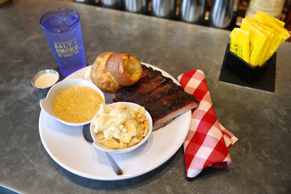 Rooftop bar at dusk with hanging string lights, people dining and lounging on curved sofas around a fire pit, a bartender at the bar, and a large arch and bridge on the city skyline.