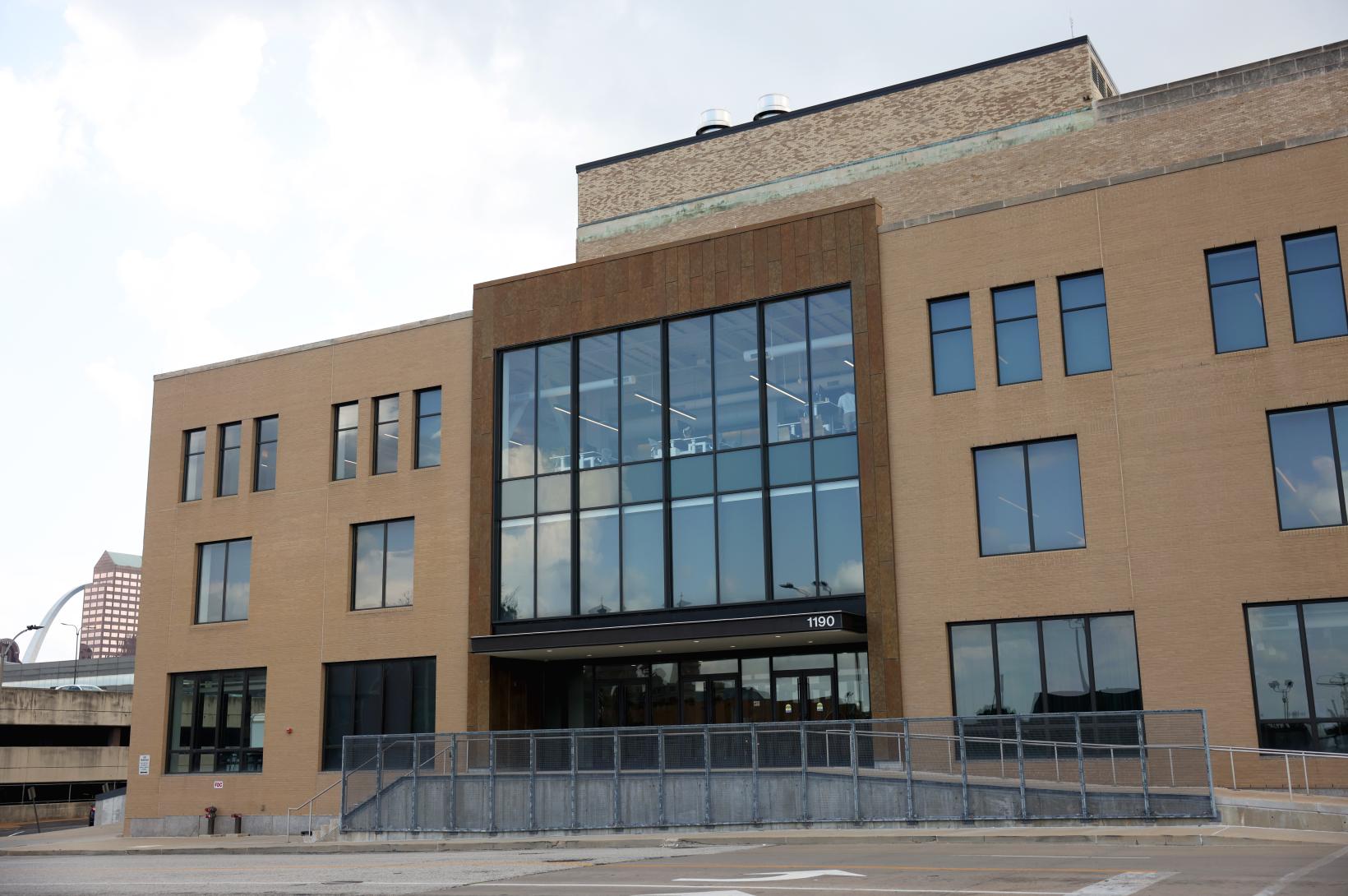 Scale-AI-Post-Building-Move-8 Three-story tan brick commercial building with a large central floor-to-ceiling glass atrium showing office lights, an accessible metal ramp leading to the main entrance marked 1190, and distant city buildings including an arch visible to the left.
