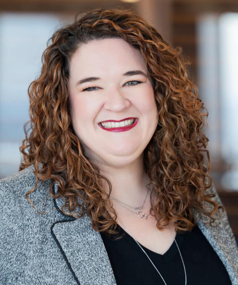 Close-up head-and-shoulders portrait of a smiling person with shoulder-length curly brown hair, wearing a gray blazer over a black top and layered necklaces, with a softly blurred indoor background.
