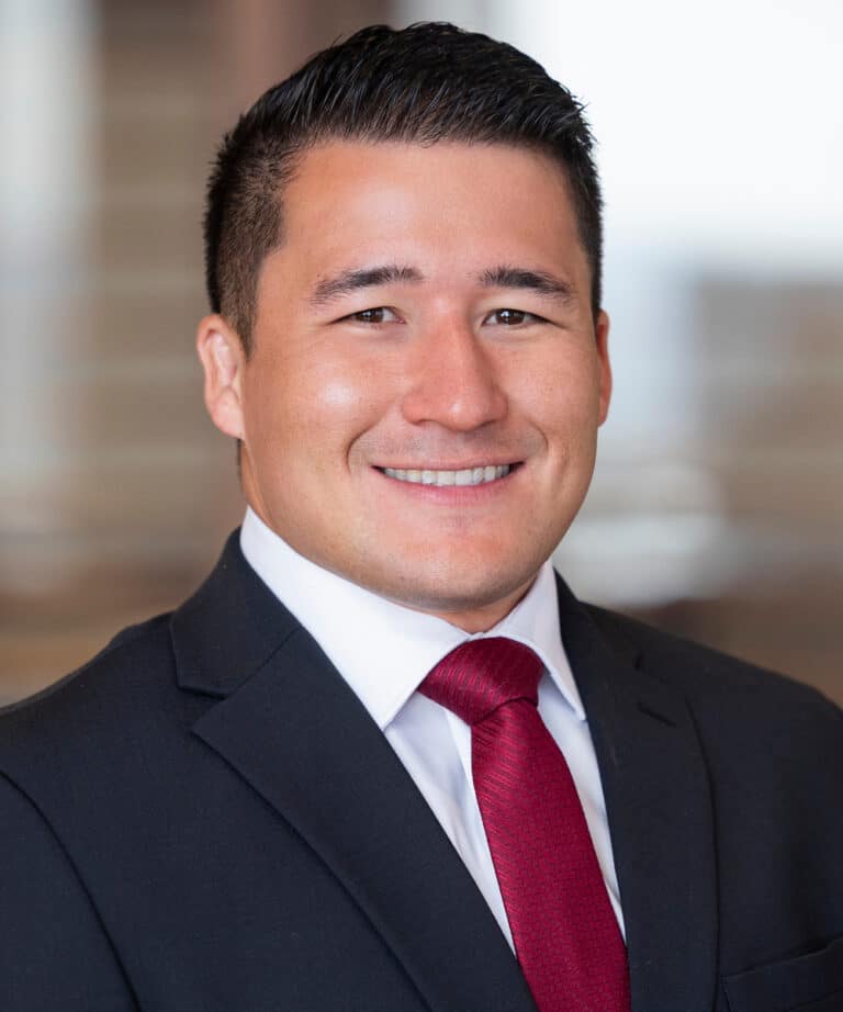 Portrait of a smiling young man with short dark hair wearing a dark suit, white shirt and red tie against a softly blurred indoor background.