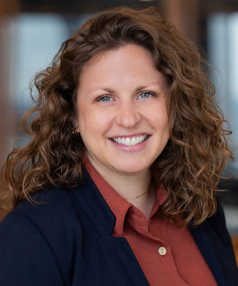 Smiling woman with curly shoulder-length brown hair and blue eyes wearing a rust-colored button-up shirt and navy blazer, photographed in a softly blurred indoor setting.