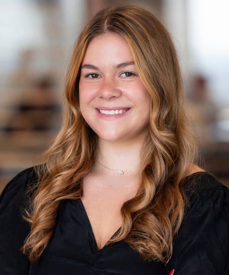Smiling woman with long wavy light-brown hair wearing a black blouse and a delicate necklace, head-and-shoulders portrait against a softly blurred indoor office background.