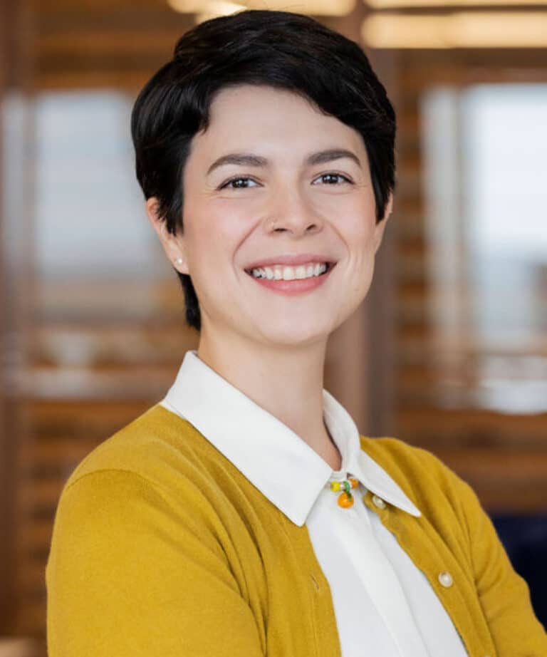 Smiling person with short dark hair wearing a mustard-yellow cardigan over a white collared shirt with a small orange pendant, standing in a softly blurred office setting.