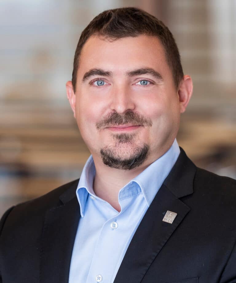 Head-and-shoulders portrait of a smiling man with short dark hair, blue eyes and a trimmed goatee, wearing a light blue button-down shirt and dark blazer with a small lapel pin against a softly blurred indoor background.
