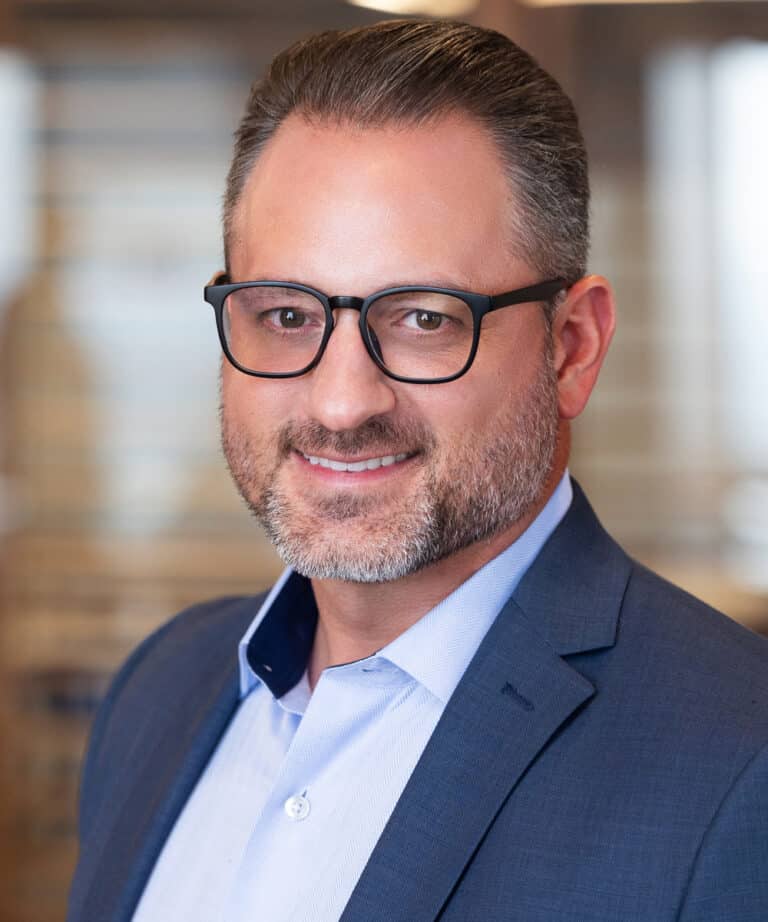 Head-and-shoulders portrait of a man with short dark hair and a salt-and-pepper beard wearing black rectangular glasses, a light blue dress shirt and navy blazer, smiling in a softly blurred office setting.