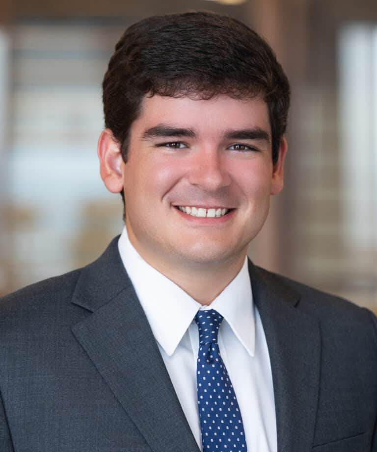 Smiling man wearing a dark gray suit, white dress shirt and blue polka-dot tie, posed against a softly blurred office background.