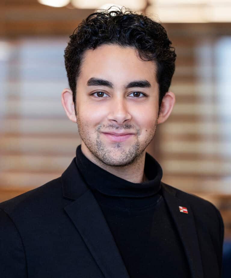 Young man with short curly dark hair and light stubble wearing a black turtleneck and blazer, smiling in front of a softly blurred interior background with horizontal wooden slats.