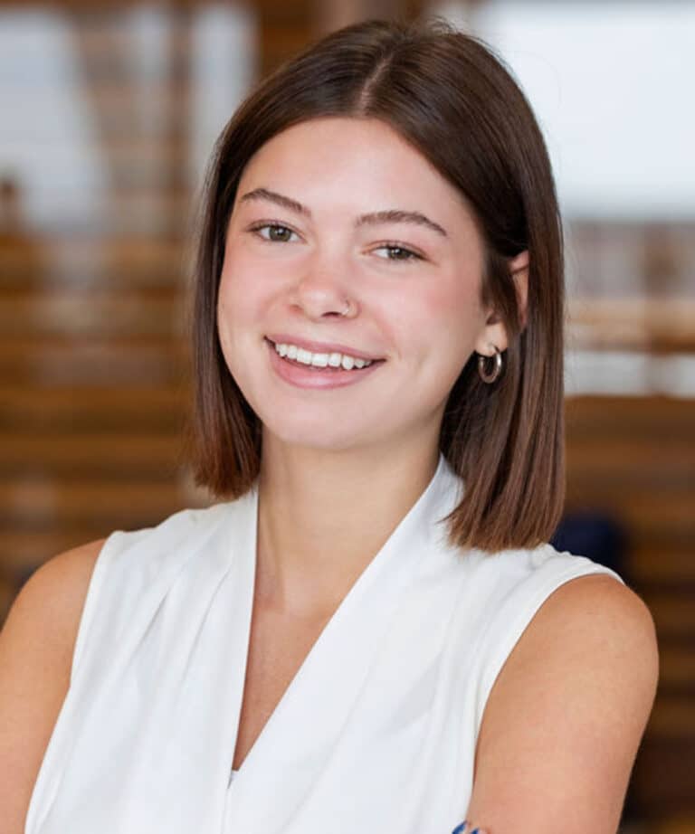 Smiling young woman with straight shoulder-length brown hair, a nose ring and small hoop earrings, wearing a white sleeveless blouse against a softly blurred wooden staircase background.