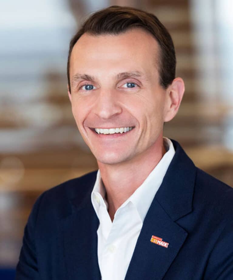 Smiling middle-aged man with short brown hair and blue eyes wearing a white dress shirt and dark blazer with a small rectangular red pin, photographed against a softly blurred indoor background.