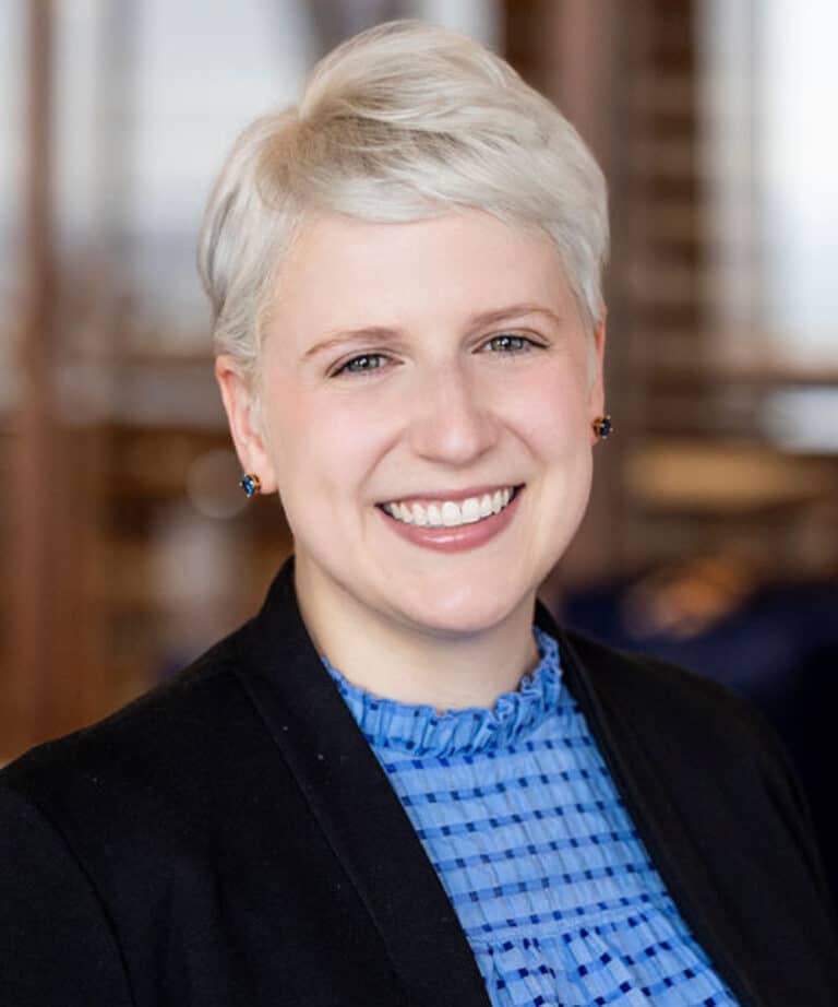 Smiling person with short platinum-blonde hair wearing small dark stud earrings, a blue patterned blouse and a black blazer, posed against a softly blurred office background.