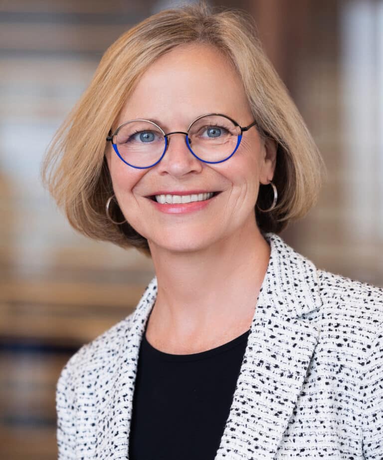 Smiling middle-aged woman with short blonde hair and round blue-rimmed glasses, wearing hoop earrings and a white textured blazer over a black top against a softly blurred indoor background.