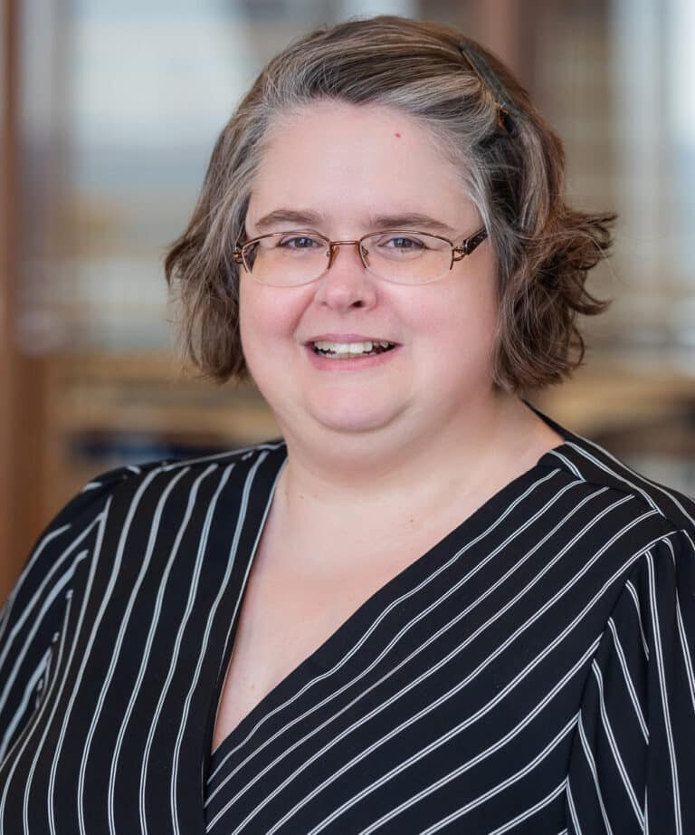 Smiling middle-aged woman with short wavy salt-and-pepper hair and glasses wearing a black V-neck blouse with white pinstripes, photographed against a softly blurred indoor background.
