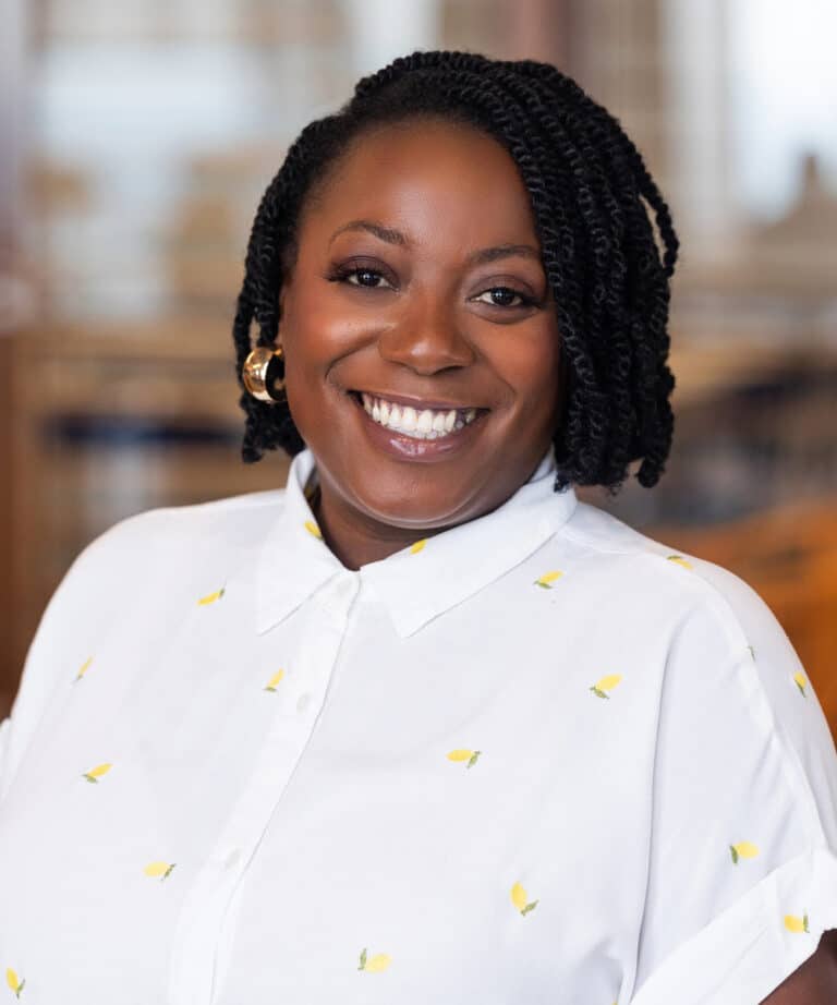 Smiling woman with short twisted hair and gold hoop earrings, wearing a white button-up shirt patterned with small yellow lemons, photographed in a softly blurred indoor setting.