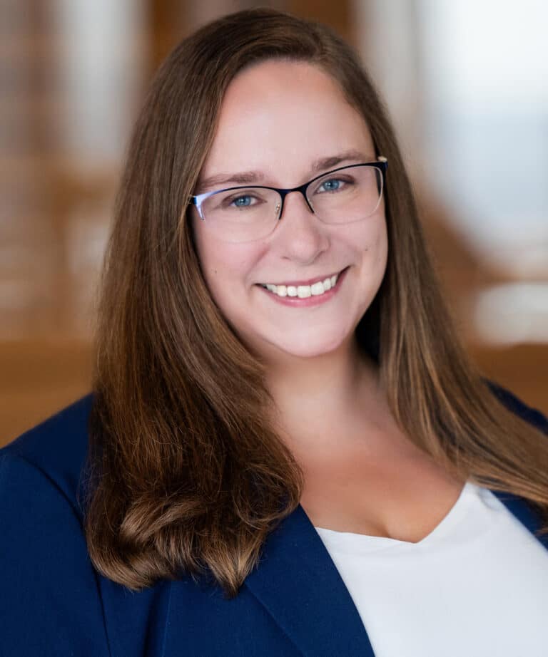 Smiling woman with long brown hair and glasses wearing a navy blazer and white top against a softly blurred indoor background.