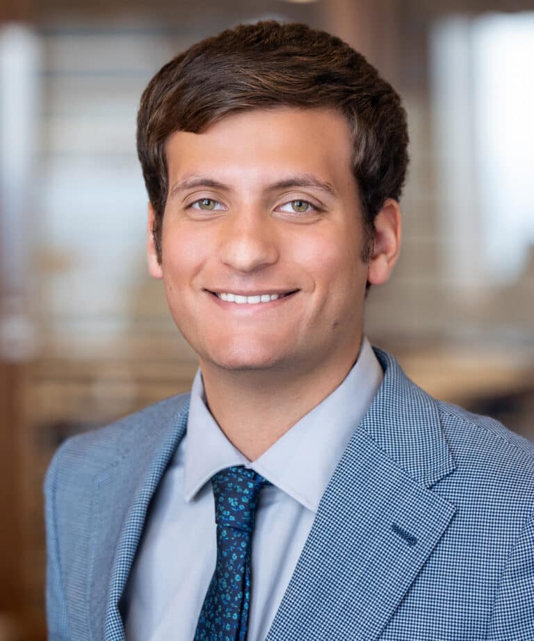 Smiling young man with short brown hair wearing a light blue checked suit jacket, pale blue shirt and dark blue patterned tie, photographed against a softly blurred office background.