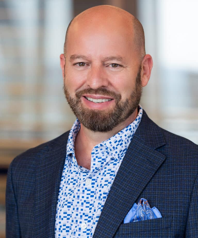 Smiling bald man with a trimmed beard wearing a white shirt with a blue square pattern, a dark checked blazer, and a blue paisley pocket square, posed against a softly blurred office background.