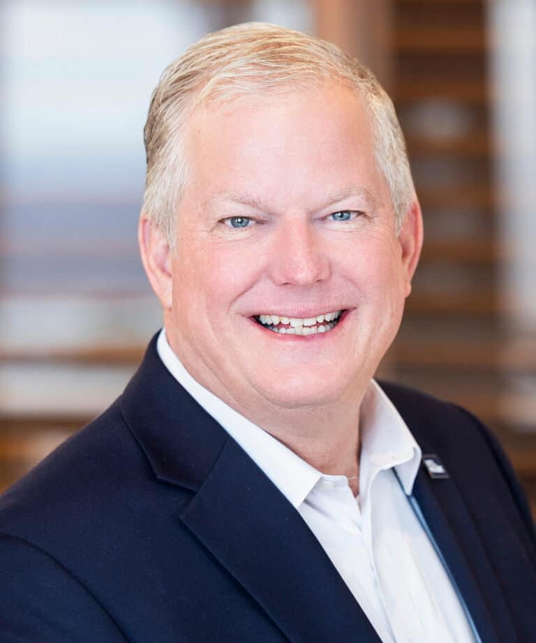 Smiling middle-aged man with short gray hair wearing a dark blazer and white dress shirt in a head-and-shoulders portrait with a softly blurred interior background.
