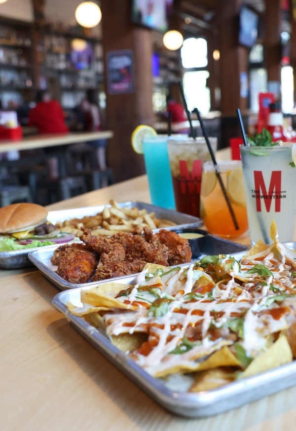Loaded nachos drizzled with sauce, with trays of fried chicken, fries and a burger and colorful cocktails on a bar.