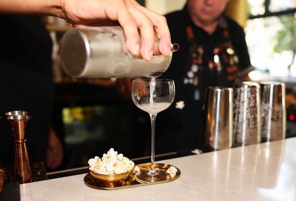 Rooftop swimming pool and sun deck with lounge chairs and a woven egg chair in the foreground, blue water bordered by a Greek-key tile band, and a city skyline under a bright, partly cloudy sky.