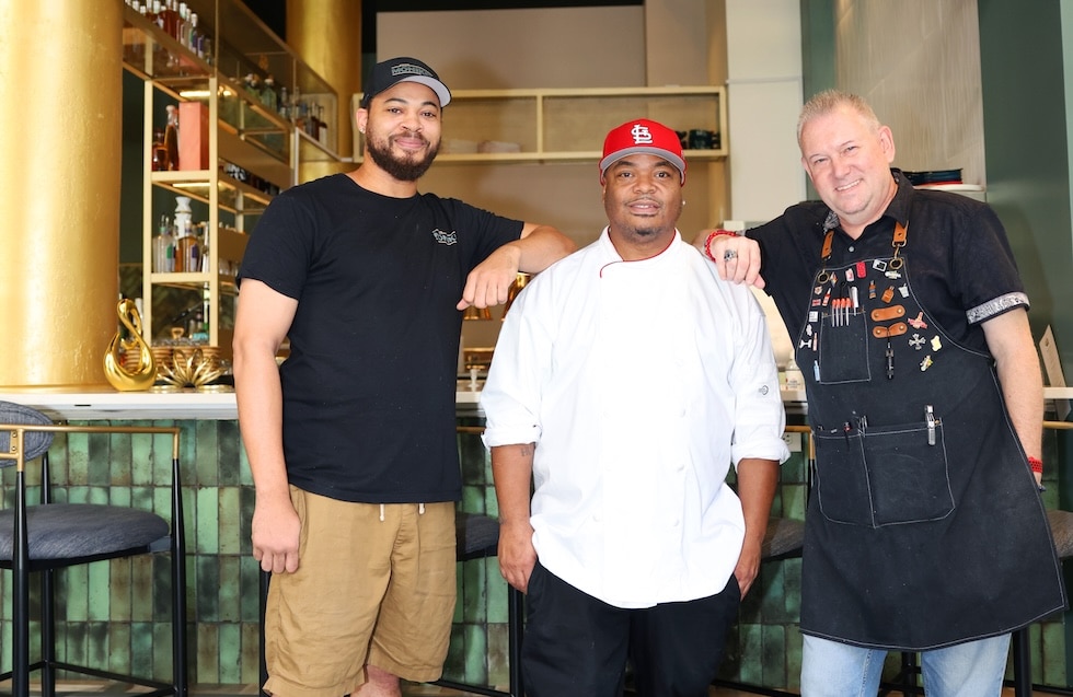 Three men—one in a black T‑shirt and cap, a chef in a white coat and red baseball cap, and a man in a black apron—stand in front of a bar.