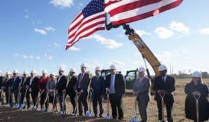 Officials in hard hats lined up with shovels at a groundbreaking under large American flags on an excavator arm.