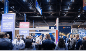 Crowded convention hall with attendees mingling around exhibition booths and overhead banners reading "STLMADE".