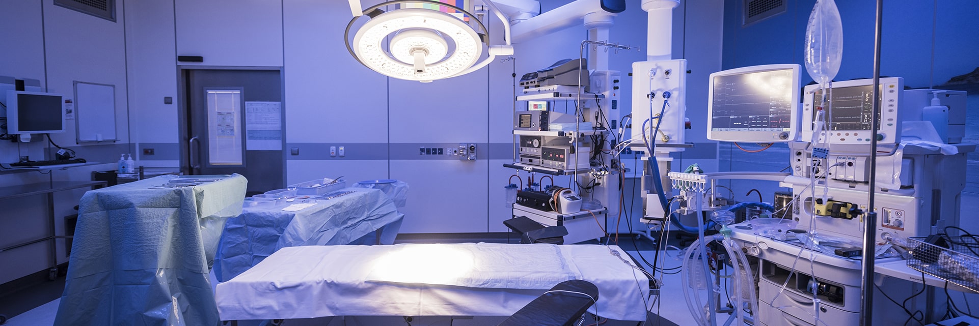 Operating room with empty surgical table under illuminated overhead lamp, monitors, anesthesia equipment and IV stand.